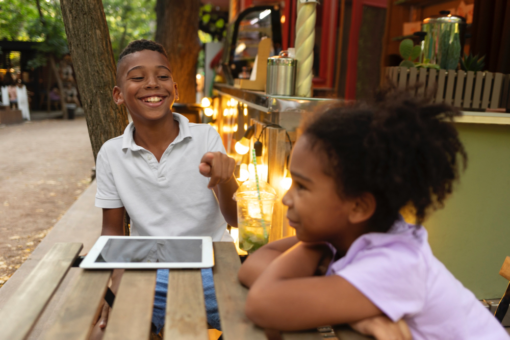 siblings laughing and with brother pointing and using iPad to talk